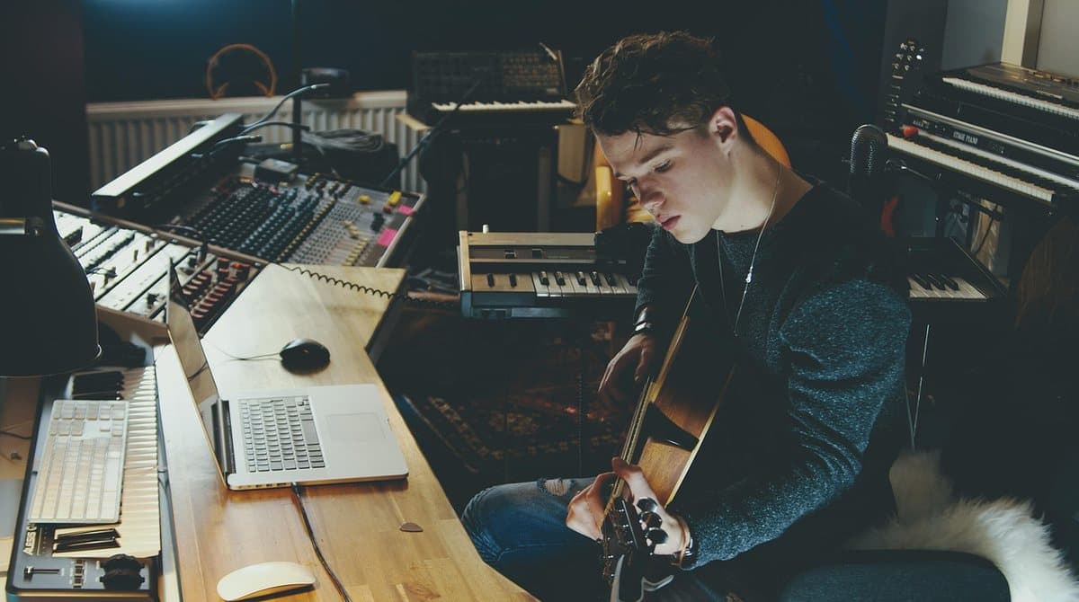 Un músico independiente tocando la guitarra en un estudio casero con portátil, mesa de mezclas y teclados