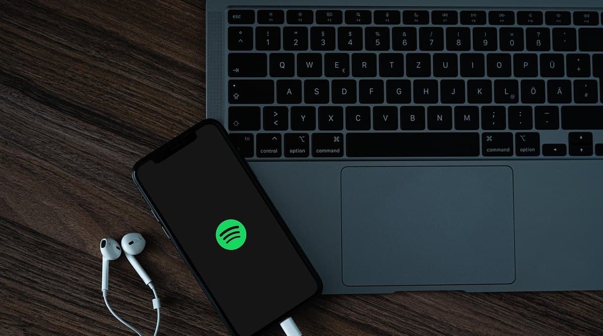 A phone showing the Spotify app resting on a laptop keyboard next to earbuds